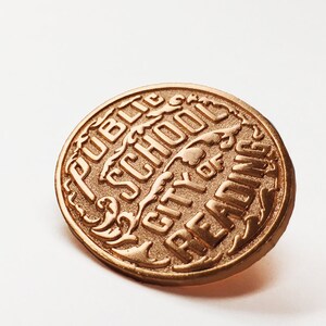 以下が含まれることがあります： 「PUBLIC SCHOOLS CITY OF READING」という文字が浮き彫りになった金色のピンバッジです。