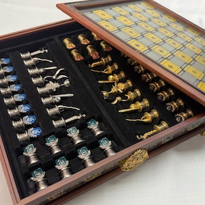 May include: A wooden chess set featuring a glass board lid and two internal velvet-lined compartments. One side contains silver-toned pieces with blue and green crests, while the other side holds gold-toned pieces with red and blue crests.