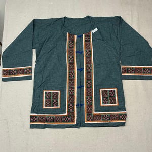 May include: A dark teal long-sleeved jacket featuring a round neckline and four blue toggle closures. The garment is accented with white-bordered panels displaying a repeating geometric pattern in red, green, and black, located on the cuffs, pockets, and front.