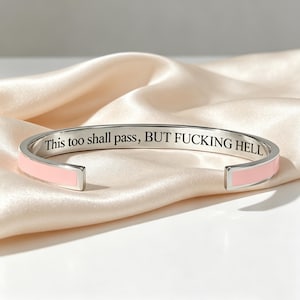 以下が含まれることがあります： 外側にライトピンクのエナメルインレイが施され、内側に「This too shall pass, BUT FUCKING HELL」という黒い刻印が入ったシルバー調のバングルブレスレット。両端が丸みを帯びたオープンデザインのブレスレットです。