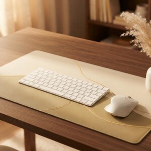 May include: A rectangular desk mat featuring a smooth, light beige and tan surface with subtle, curved line patterns. A white wireless keyboard and a matching white computer mouse are placed on the mat, alongside a small, speckled ceramic vase.