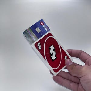 Puede incluir: Un tarjetero rectangular con un diseño de carta de reverso de Uno en color rojo, flechas blancas y un acabado brillante. El tarjetero está parcialmente abierto, dejando ver una tarjeta de crédito azul de US Bank con chip plateado y símbolo contactless.