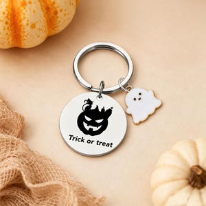 Könnte beinhalten: Ein runder Schlüsselanhänger aus silberfarbenem Metall mit einer schwarzen Gravur eines Kürbisses, einer Katze und eines Grabsteins, ergänzt durch den Schriftzug "Trick or treat" und einen kleinen weißen Geisteranhänger aus Emaille.