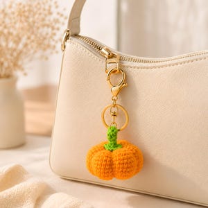 May include: A crochet pumpkin keyring featuring a bright orange pumpkin with a green stem, attached to a gold-toned metal lobster clasp and a circular ring. The keyring is clipped onto the zip pull of a cream-coloured shoulder bag.