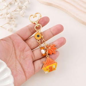 May include: A gold-toned metal keyring featuring a heart-shaped clasp, a sunflower charm, a white pearl bead, an orange maple leaf charm, and a house-shaped charm with an orange roof and yellow walls, all suspended from a delicate link chain.