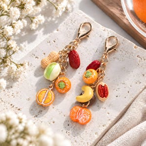 May include: Two gold-tone metal keychains featuring miniature charms of various fruits and nuts, including a peanut, pistachio, persimmon, orange slice, cashew, and red date, all attached to a sturdy lobster clasp with a linked chain design.