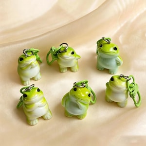 May include: Six small, light green and white frog figurines, each attached to a silver-toned metal lobster clasp keychain. The frogs are depicted in various standing poses, featuring smooth surfaces, dark eyes, and a glossy finish on their bodies.