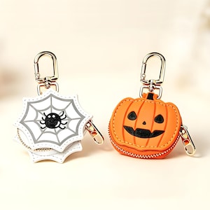 May include: Two small zippered pouches with gold-tone metal lobster clasps. One pouch is white and shaped like a spider web with a black spider graphic. The other pouch is orange and shaped like a jack-o'-lantern with a black face design.