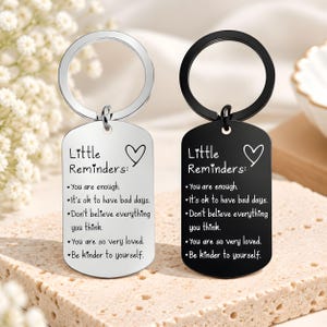 May include: Two rectangular metal keychains, one silver and one black, each featuring a ring at the top. Both tags are engraved with the text "Little Reminders:" followed by five positive affirmations and a heart symbol, all in a clear font.