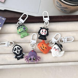 May include: A collection of six Halloween-themed keychains featuring colorful plastic charms attached to white lobster clasps. The charms include a green Frankenstein monster, a black-hooded figure, a ghost, a pumpkin, a purple spider, and a ghost.