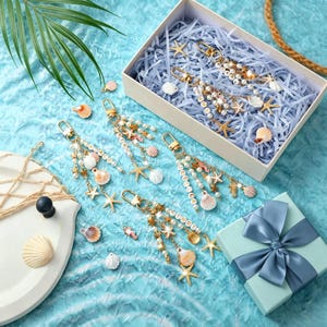 May include: Four gold-toned keychains featuring white beads, small seashells, and starfish charms. Some beads are lettered, spelling names like CINDY. The keychains are arranged on a blue textured surface, with some inside a white gift box.