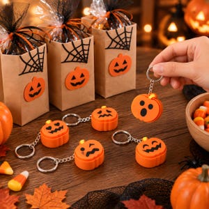 Puede incluir: Una colección de llaveros de calabaza naranja con varias expresiones faciales negras, incluyendo ojos de corazón, un ojo guiñando y una boca sorprendida. Cada llavero cuenta con una anilla y una cadena de metal plateado.