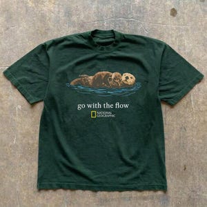 Może przedstawiać: Ciemnozielony t-shirt z krótkim rękawem z grafiką brązowej wydry morskiej unoszącej się w niebieskiej wodzie. Poniżej wydry znajduje się biały napis "go with the flow" oraz żółto-białe logo National Geographic.