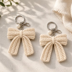 May include: Two cream-colored crochet bow keychains, each featuring a textured yarn bow attached to a silver-toned metal split ring and a lobster clasp, accented with a small, round, off-white bead hanging from the chain link connector.