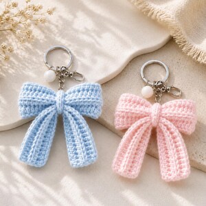May include: Two crochet bow keychains, one in light blue and one in soft pink. Each bow features a silver-toned metal keyring, a small lobster clasp, and a single white spherical bead attached to the chain link above the fabric bow structure.