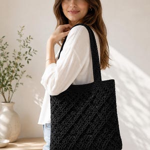 May include: A black crochet tote bag featuring a repeating diamond-shaped floral pattern throughout the body. The bag has two long, sturdy shoulder straps made of the same black yarn, providing a consistent texture and color across the entire piece.