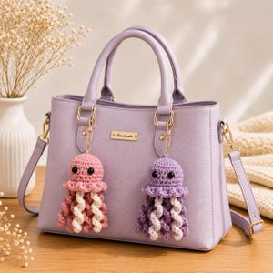 May include: A lavender handbag with two top handles and a detachable shoulder strap. The bag features a gold-toned metal plaque with the word Handmade and two crochet jellyfish charms in pink and purple hanging from the gold hardware rings.
