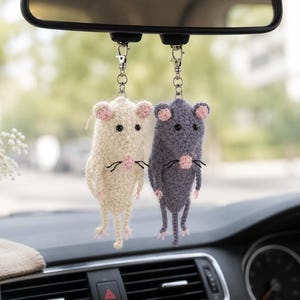 Könnte beinhalten: Zwei gehäkelte Ratten-Ornamente, die am Rückspiegel eines Autos hängen. Eine Ratte ist cremefarben, die andere dunkelgrau. Beide haben kleine rosa Ohren, eine rosa Nase, schwarze Perlenaugen und dünne schwarze Schnurrhaare an silbernen Metallclips.