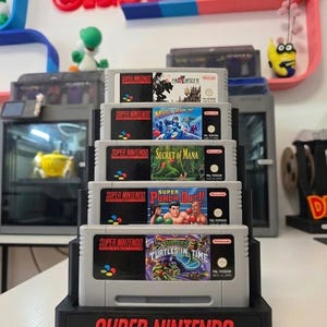 Könnte beinhalten: Ein schwarzer Kunststoff-Präsentationsständer mit fünf vertikal gestapelten grauen Super Nintendo-Spielkassetten. Der Ständer trägt an der Basis den roten Schriftzug Super Nintendo Entertainment System. Die Kassetten umfassen Final Fantasy III, Mega Man X, Secret of Mana, Super Punch-Out!! und Teenage Mutant Ninja Turtles: Turtles in Time.