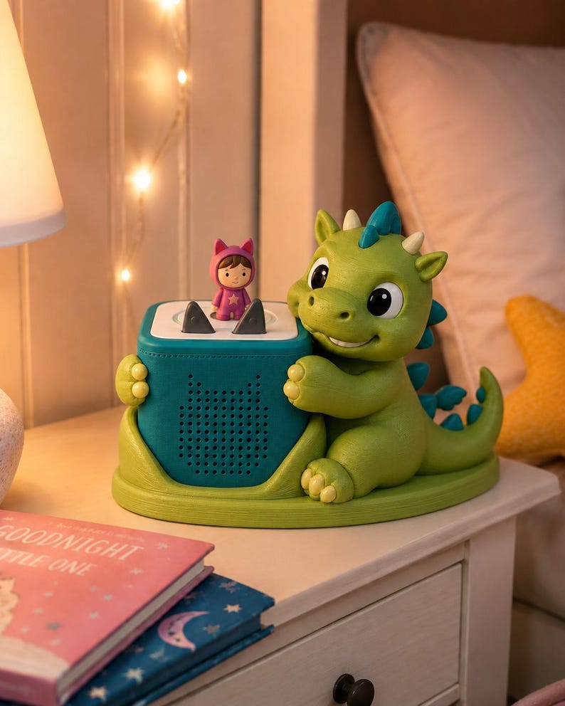 Puede incluir: Una figura de drag&oacute;n verde que sostiene una base de altavoz cuadrada de color verde azulado con una superficie superior blanca. Un peque&ntilde;o personaje con un traje rosa con capucha se encuentra sobre el altavoz. El altavoz tiene un panel frontal perforado.