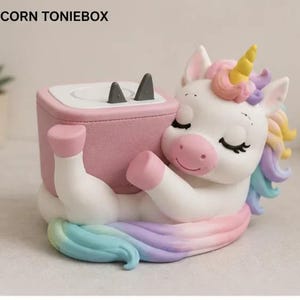 May include: A white unicorn figure with a yellow horn and pastel rainbow mane, hugging a pink square speaker box labelled UNICORN TONIEBOX. The speaker has a white top surface with two small grey triangular ears protruding from the top edge.