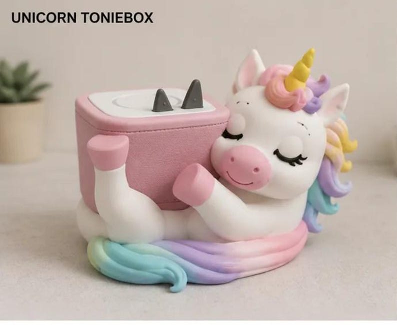 May include: A white unicorn figure with a yellow horn and pastel rainbow mane, hugging a pink square speaker box labelled UNICORN TONIEBOX. The speaker has a white top surface with two small grey triangular ears protruding from the top edge.