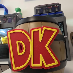 Puede incluir: Un barril cilíndrico con cuerpo color canela y bandas negras, que presenta un emblema grande y prominente en rojo y amarillo con las letras DK adheridas al frente. El objeto tiene un acabado mate liso y un diseño de paneles verticales.