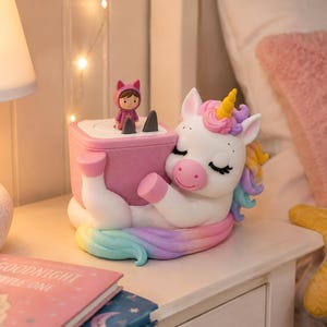 May include: A white unicorn night light with a pastel rainbow mane and tail, featuring closed eyes and a yellow horn. The unicorn holds a pink square base with a small figure wearing a purple cat-ear hood standing on top of the flat surface.