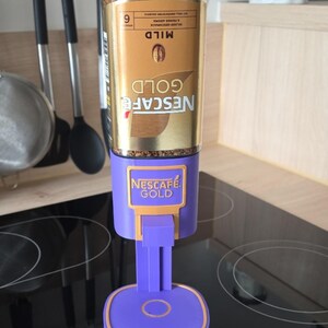 May include: A Nescaf&eacute; Gold instant coffee jar mounted upside down into a purple plastic dispenser stand. The jar features a gold label with the text Nescaf&eacute; Gold and Mild. The purple stand has a square base with a gold trim and a lever mechanism.