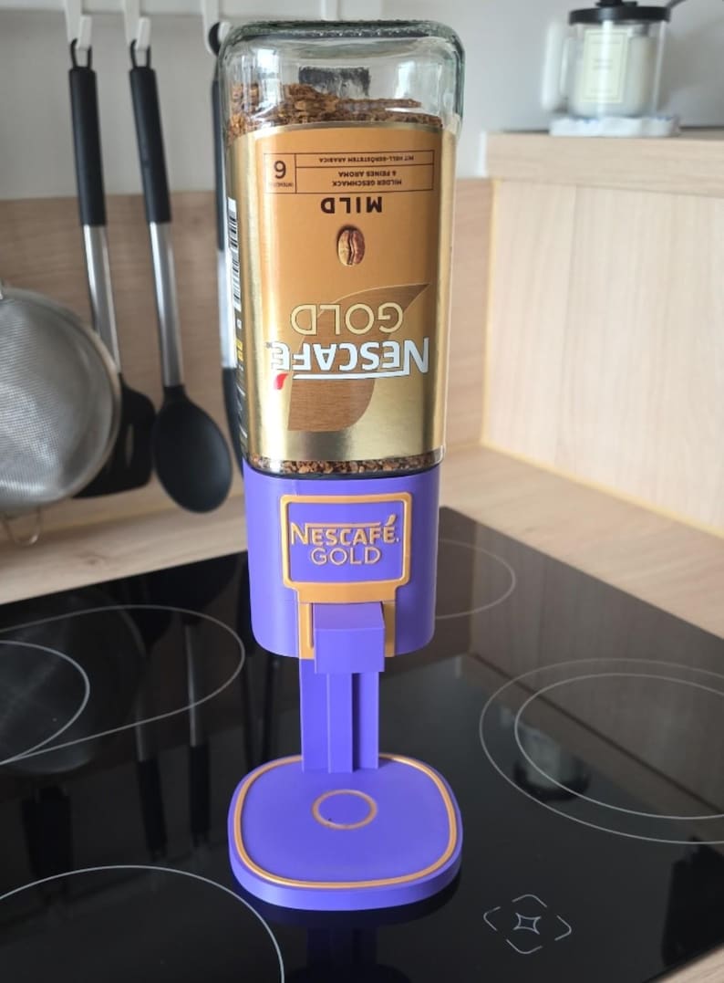 May include: A Nescaf&eacute; Gold instant coffee jar mounted upside down into a purple plastic dispenser stand. The jar features a gold label with the text Nescaf&eacute; Gold and Mild. The purple stand has a square base with a gold trim and a lever mechanism.