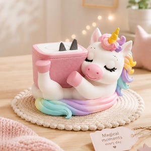 May include: A white unicorn figurine with a yellow horn and a pastel rainbow mane, hugging a pink square box with two small black triangular accents on top. A light pink tag attached to the base features the text Magical moments every day.