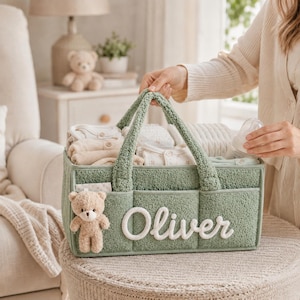 May include: A sage green textured fabric diaper caddy with two handles, featuring the name Oliver in white script lettering on the front and a small beige plush teddy bear attached to the side, filled with folded baby clothing and accessories.