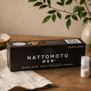以下が含まれることがあります： NATTOMOTOと書かれた黒い長方形の箱。Bacillus subtilis strain Takahashi ver.natto、Made in Japanの白い文字とQRコードが印刷されています。白い粉末が入った小さな透明なプラスチック製の小瓶が、箱の横の布の上に置かれています。