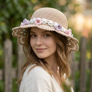 May include: A beige crochet sun hat featuring a wide, scalloped brim and a decorative band of white crocheted trim. The hat is adorned with small, hand-crocheted flowers in shades of soft pink and light purple arranged around the crown.
