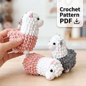 May include: Three crocheted alpaca figures featuring textured yarn loops. One is white and pink, another is white and gray, and the third is pink and white. Each has small black bead eyes and pink stitched noses. Text reads: Crochet Pattern PDF.