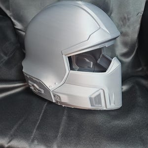 May include: A silver-coloured full-face helmet featuring a smooth, rounded dome, a wide horizontal visor opening, and angular geometric detailing along the lower jaw and side panels. The helmet has a solid, moulded construction with a matte finish.