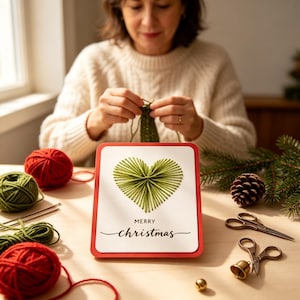 Könnte beinhalten: Eine rechteckige Grußkarte mit rotem Rand, die ein grünes Herz aus Fadengrafik und den schwarzen Schriftzug Merry Christmas zeigt. Die Karte ist umgeben von roten und grünen Wollknäueln, einer Metallschere und einem kleinen Tannenzapfen.
