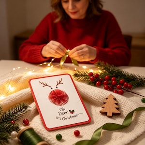 Puede incluir: Una tarjeta de felicitación rectangular blanca con borde rojo que presenta una ilustración de reno con una nariz de arte en hilo rojo y astas dibujadas en tinta marrón. El texto dice Oh Deer! It's Christmas con un pequeño gráfico de acebo.