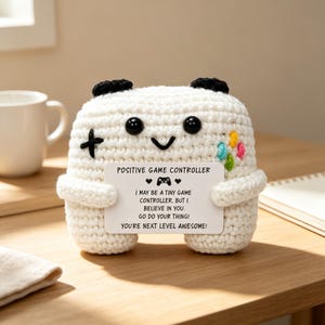 May include: A white crocheted plush toy shaped like a game controller with black eyes, a smiling mouth, and two black top accents. It holds a white card that reads: POSITIVE GAME CONTROLLER, I MAY BE A TINY GAME CONTROLLER, BUT I BELIEVE IN YOU.