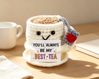 May include: A cream-colored crocheted mug with a brown top, black eyes, and a smiling mouth. It holds a white card with the text "YOU'LL ALWAYS BE MY BEST-TEA" and features a grey crocheted tea bag tag with a red heart attached to the side.