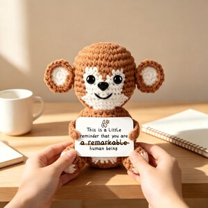 Puede incluir: Un peluche de mono tejido a ganchillo en color marrón y blanco que sostiene una tarjeta rectangular blanca con el texto: This is a Little reminder that you are a remarkable human being. El mono tiene ojos negros grandes, nariz negra y orejas redondas con centros blancos.