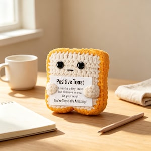 May include: A crocheted toast figure with a white center and orange crust, featuring black plastic eyes and a small mouth. It holds a white card that reads: Positive Toast, I may be a tiny toast but I believe in you. Go your way! You're Toast-ally Amazing!
