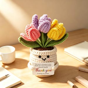 May include: A crocheted potted plant featuring pink, purple, and yellow tulips with green leaves. The cream-colored pot has a smiling face and holds a white sign that reads: The world is fortunate to have people who make things better wherever they go thank you for being one of them.