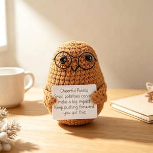 Könnte beinhalten: Eine braune, gehäkelte Kartoffelfigur mit einer schwarzen Drahtbrille. Die Figur hält ein weißes, rechteckiges Schild mit der Aufschrift: Cheerful Potato, Small potatoes can show make a big impact! Keep pushing forward, you got this!