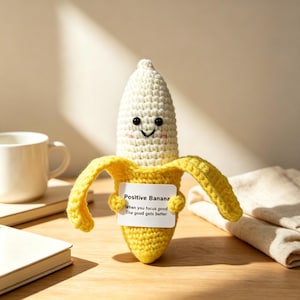 Puede incluir: Un peluche de banana tejido a ganchillo con la parte superior blanca y la cáscara amarilla. Tiene ojos de cuentas negras, una sonrisa bordada y mejillas rosadas. La banana sostiene una tarjeta rectangular blanca con el texto: Positive Banana. When you focus good, The good gets better.