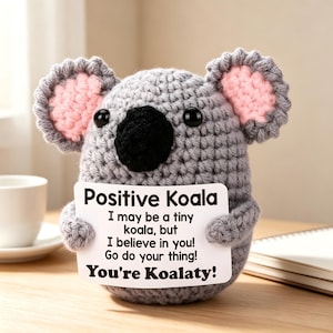May include: A grey crocheted koala plush toy with pink inner ears and a black nose, holding a white sign that reads: Positive Koala, I may be a tiny koala, but I believe in you! Go do your thing! You're Koalaty! The plush has two black bead eyes.