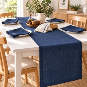 May include: A long, rectangular navy blue table runner with a textured weave, paired with four matching navy blue cloth napkins. The runner is centred on a white table, creating a clean, uniform look for dining arrangements and table settings.