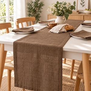 Peut inclure: Un long chemin de table rectangulaire de couleur marron, fabriqué dans un tissu texturé et posé à plat sur une table à manger blanche. Il est accompagné de serviettes en tissu marron assorties, pliées sur des assiettes blanches.