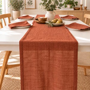 Peut inclure: Un long chemin de table rectangulaire en lin couleur rouille centré sur une table blanche. Quatre serviettes en tissu assorties sont pliées sur des assiettes blanches. Un pichet en céramique blanche contient des végétaux et un bol en bois des poires.