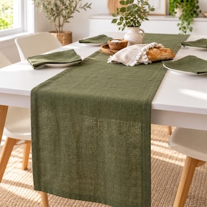 Puede incluir: Un camino de mesa largo y rectangular de color verde oliva con una textura tejida visible, centrado en una mesa de comedor blanca. El camino de mesa se acompaña de servilletas de tela verde oliva a juego dobladas sobre platos blancos.